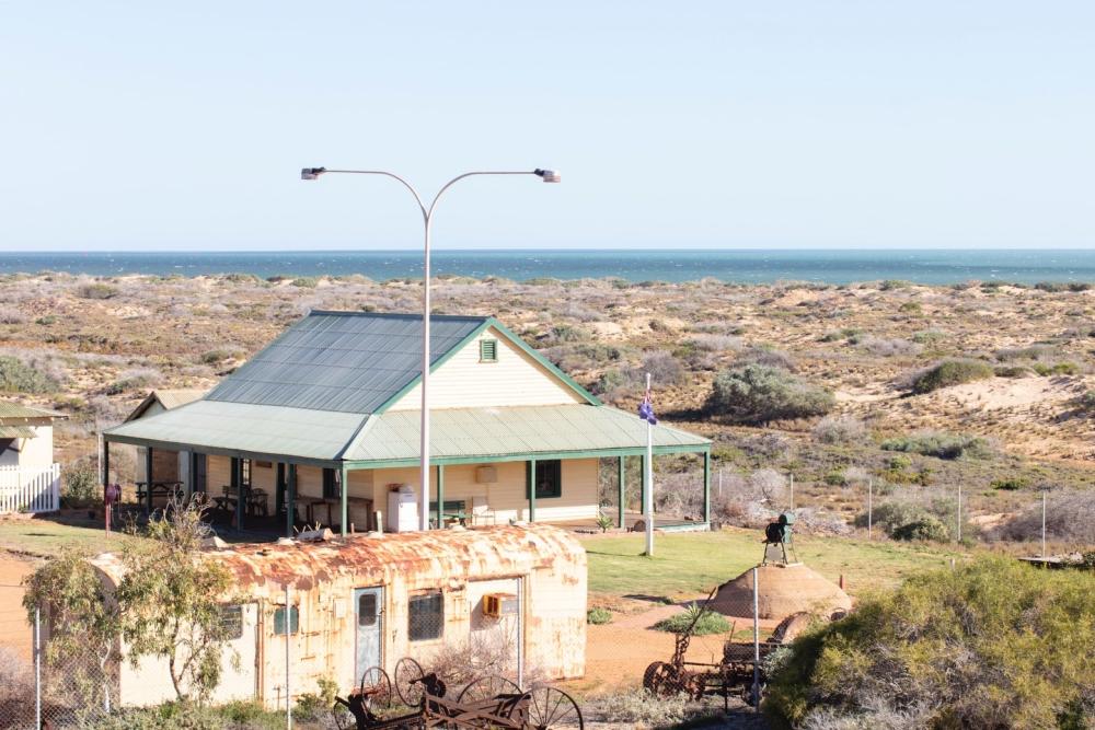 white cottage with green verandah surrounded by shrub land with the ocean viable in the background.